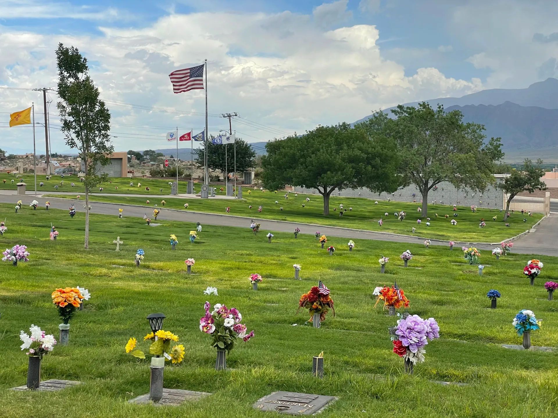 Cemetery with green grass, colorful flowers, and flags waving under a cloudy sky.