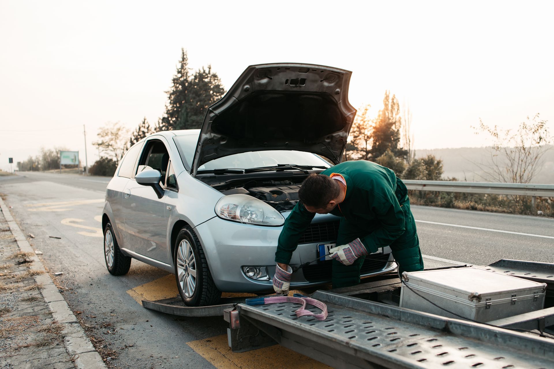 Tow truck operator securing a broken-down car onto a flatbed on a roadside.