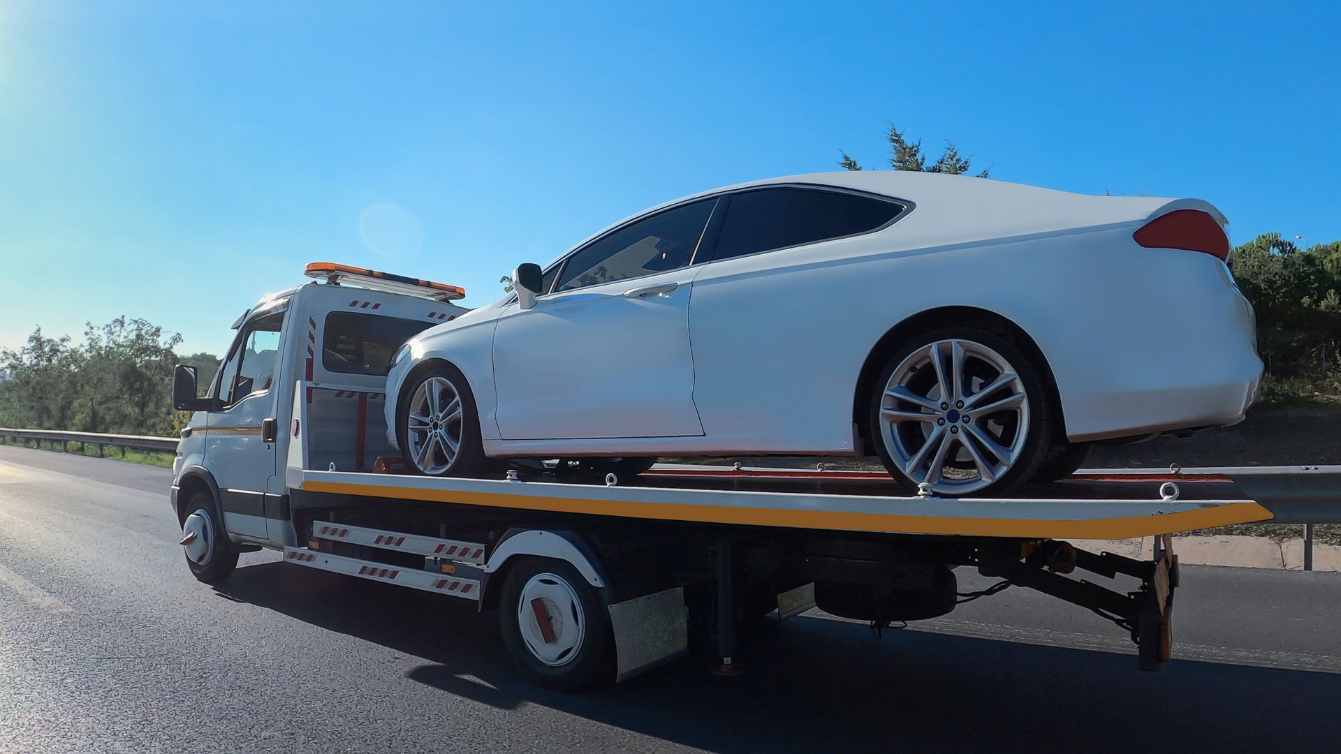A white sports car on a tow truck driving down a highway in daylight.]