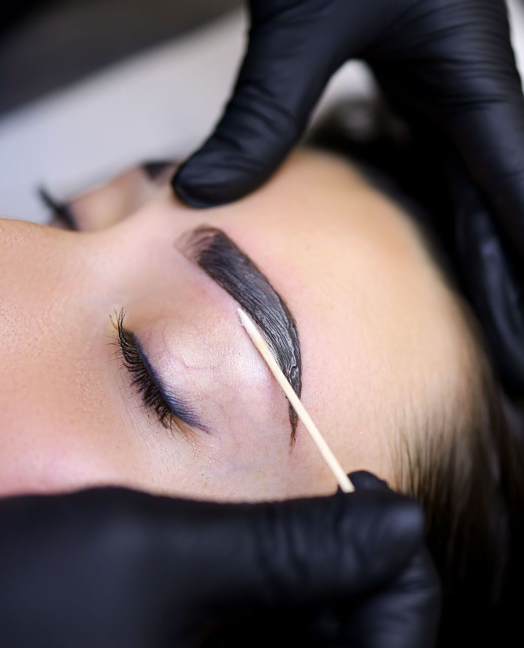 Person receiving eyebrow microblading; black gloved hands, close-up, focused.