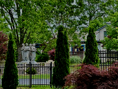 A fenced garden with two stone statues, tall green trees, and shrubs in front of a house.