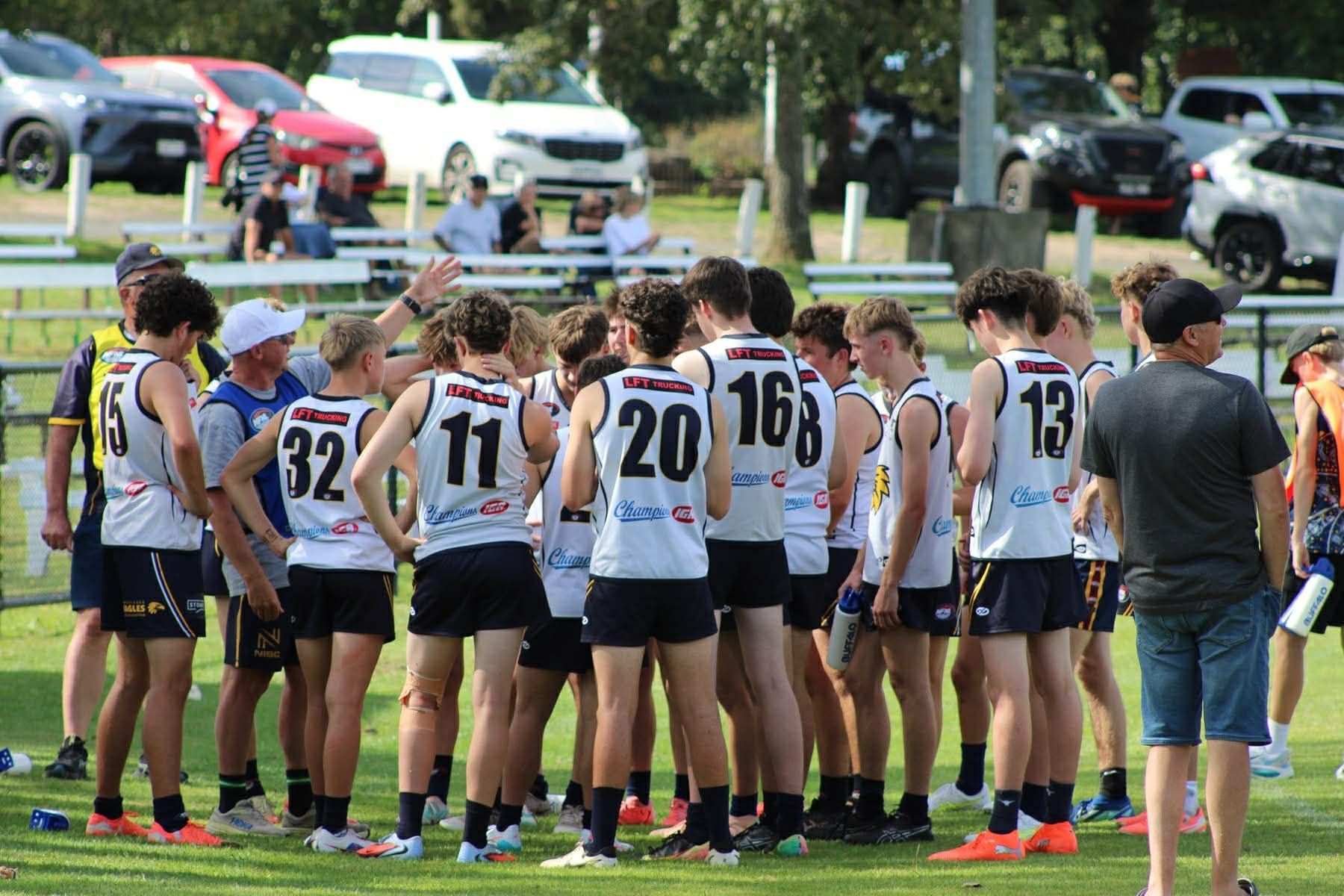 Athletes in white jerseys huddle on a grassy field during a sports match, with cars and spectators in the background