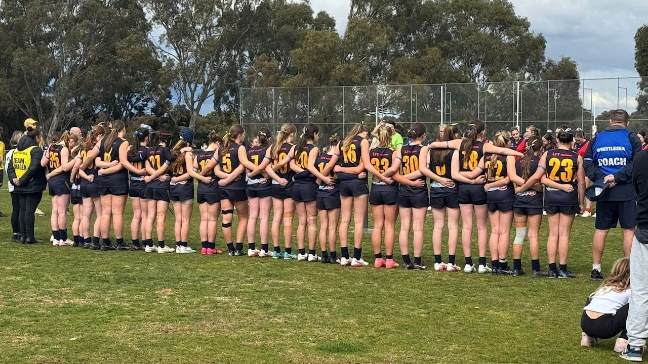 Sports team lined up on a grassy field, facing away, with a coach in blue at right.