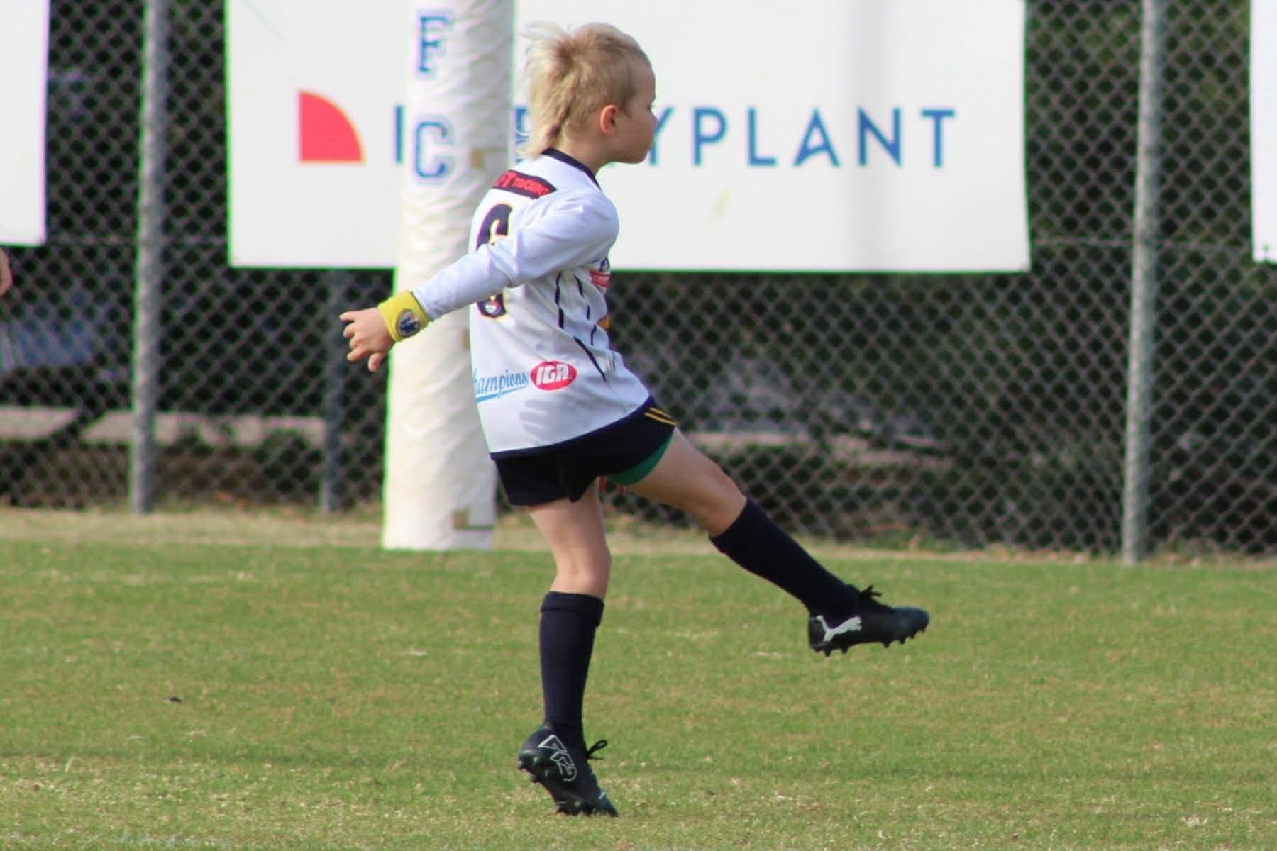 Soccer player in gray jersey kicks a ball on a grassy field near a chain-link fence