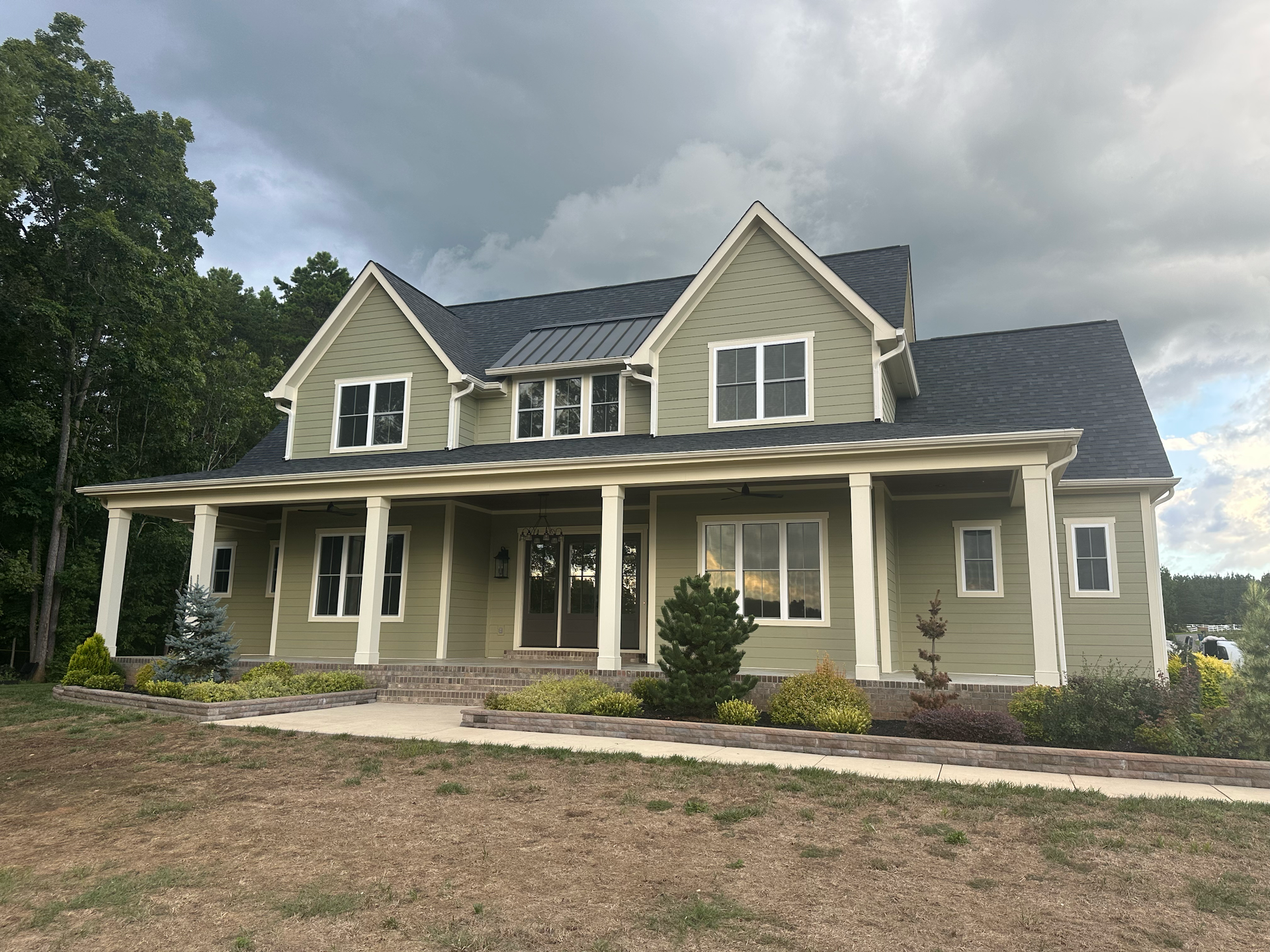 Green two-story house with a wrap-around porch under a cloudy sky.