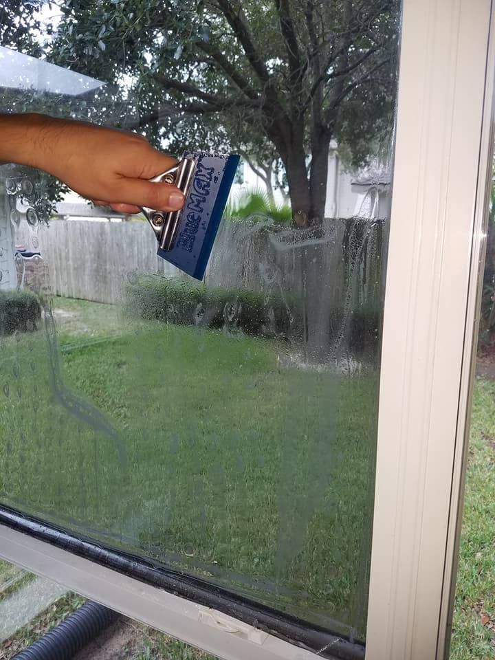 Hand using a blue squeegee on a window, outside scene of green grass and fence.