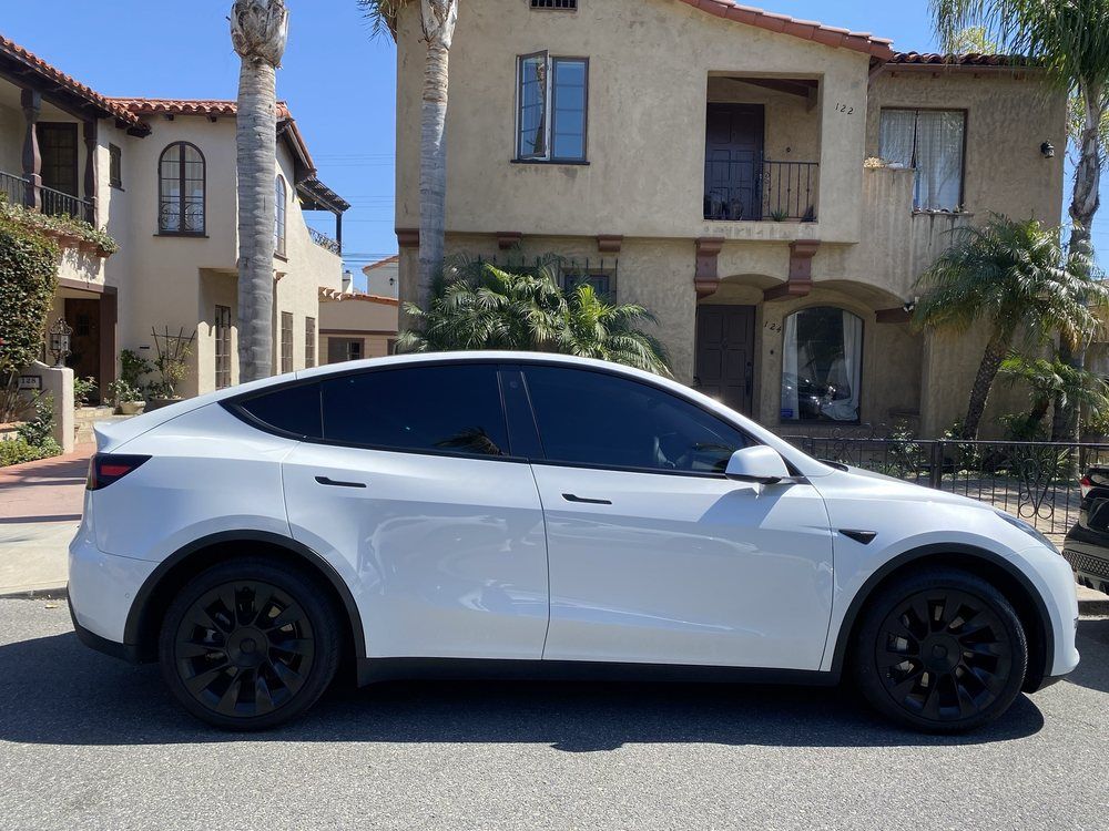 White Tesla Y parked on a street with houses in the background; sunny day.