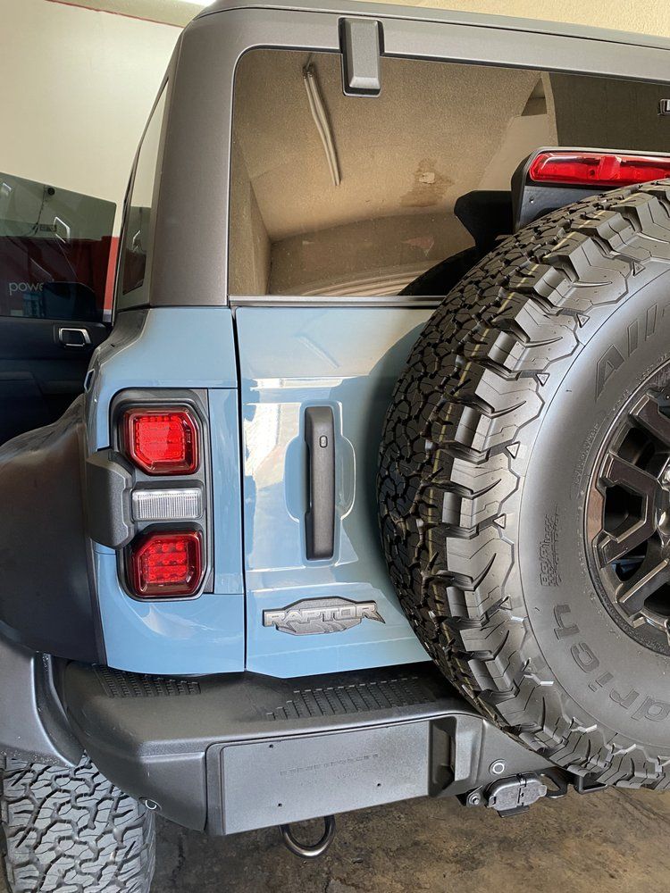 Rear view of a light blue Ford Bronco SUV with a spare tire. Black trim and taillights are visible.