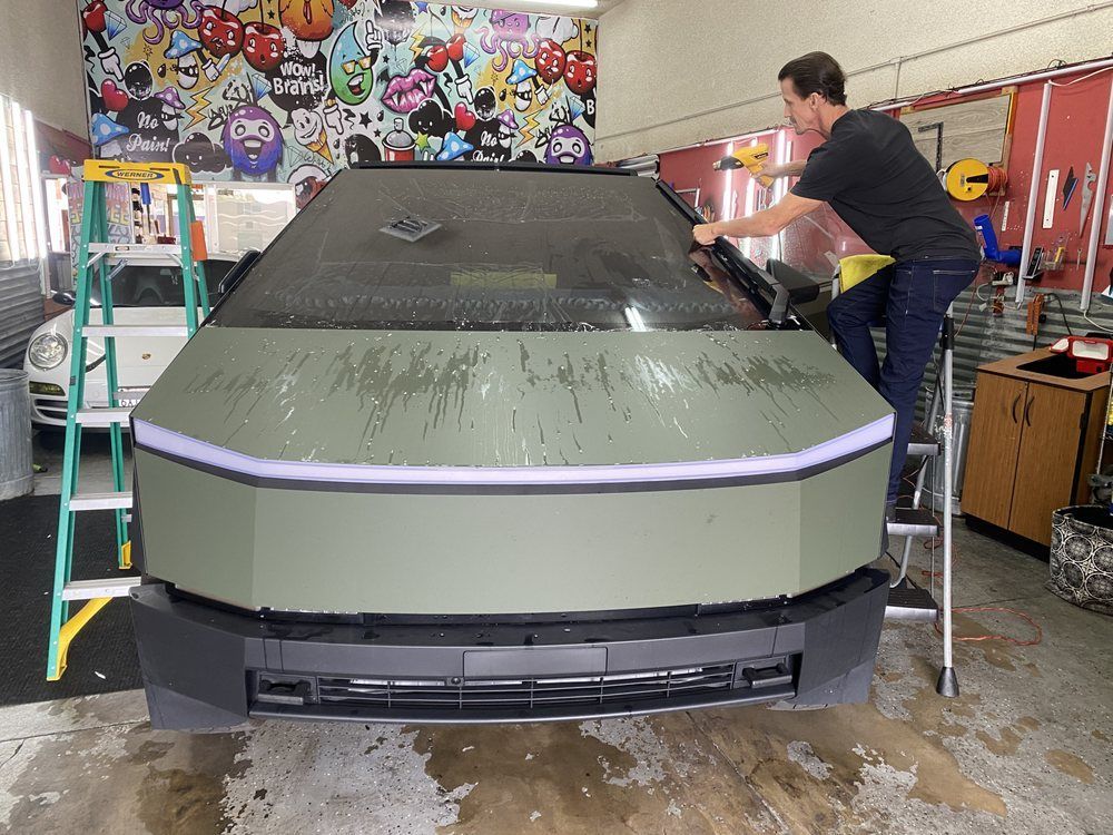 A man applies film to the windshield of a green Tesla Cybertruck in a garage.