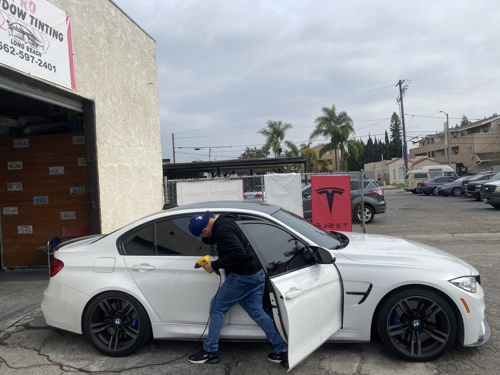 Man applying window tint to a white BMW sedan outside a shop.