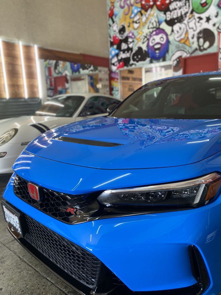Bright blue Honda Civic Type R with a red badge, parked inside a garage next to a white Porsche.