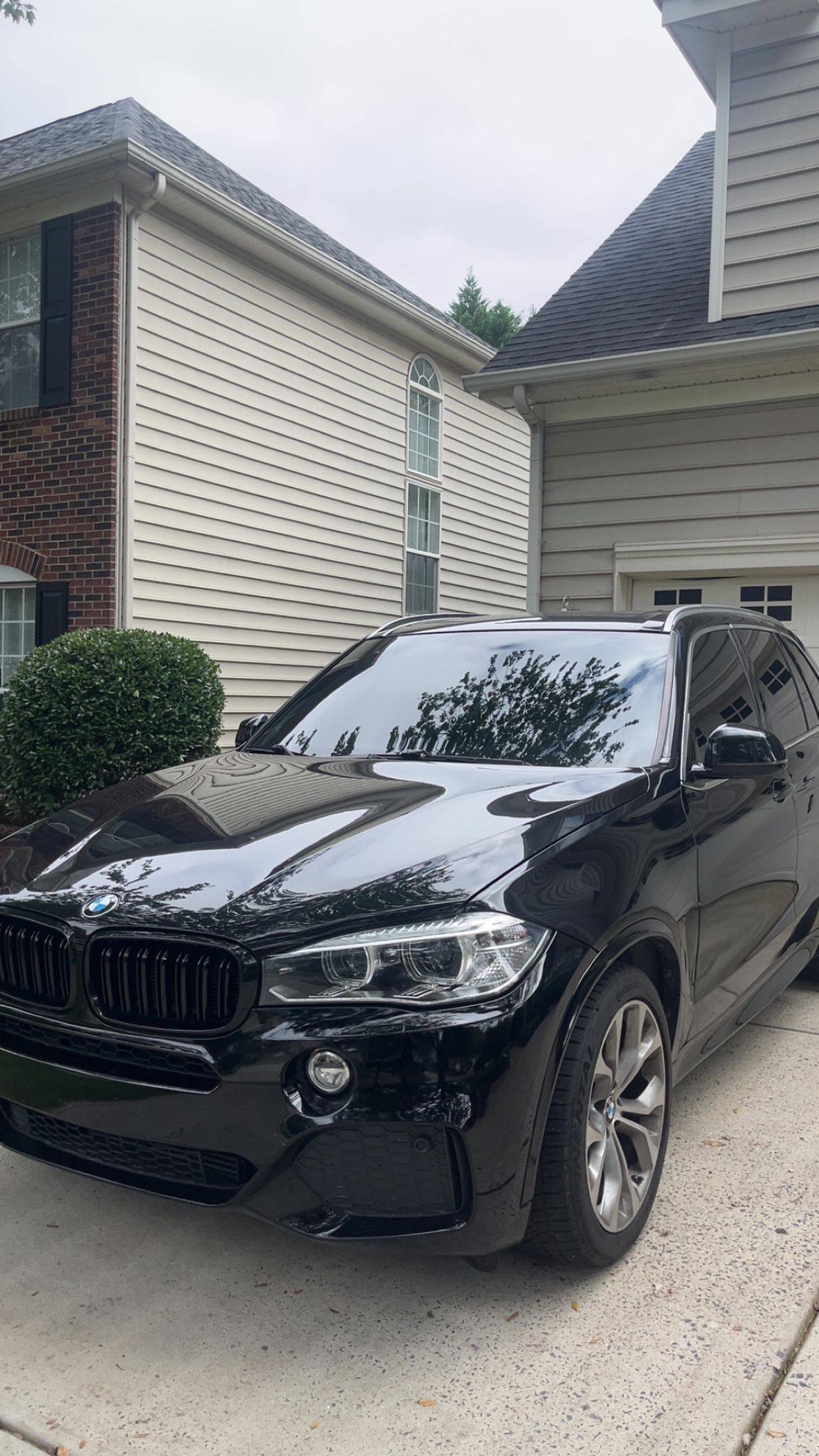 Black BMW X5 SUV parked outside a house with tan siding. Overcast sky.
