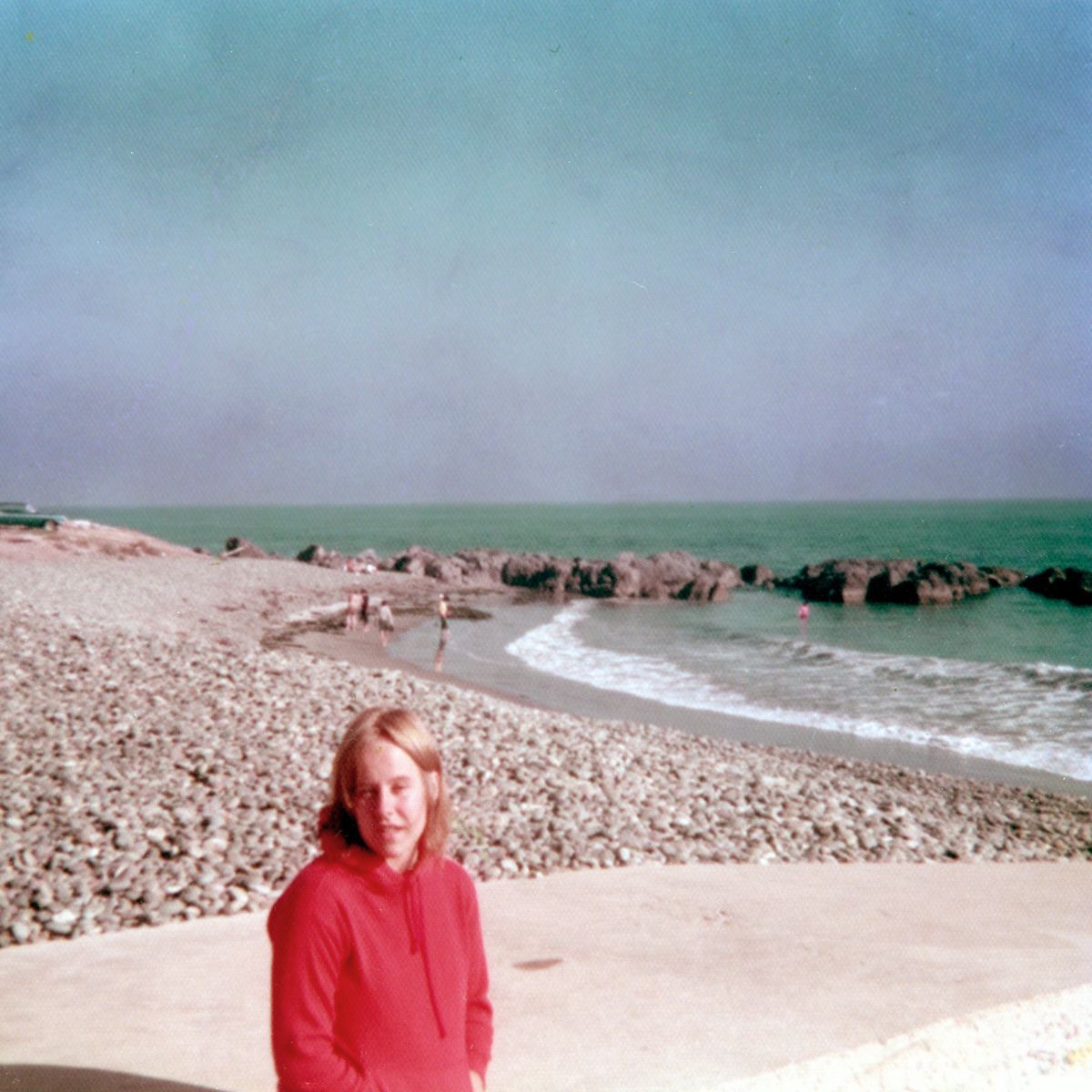 A woman in a red shirt stands on a rocky beach near the ocean