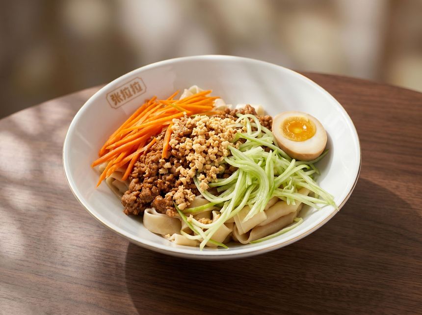 Bowl of mixed noodles with shredded vegetables, minced meat, and a halved soft-boiled egg on a wooden table