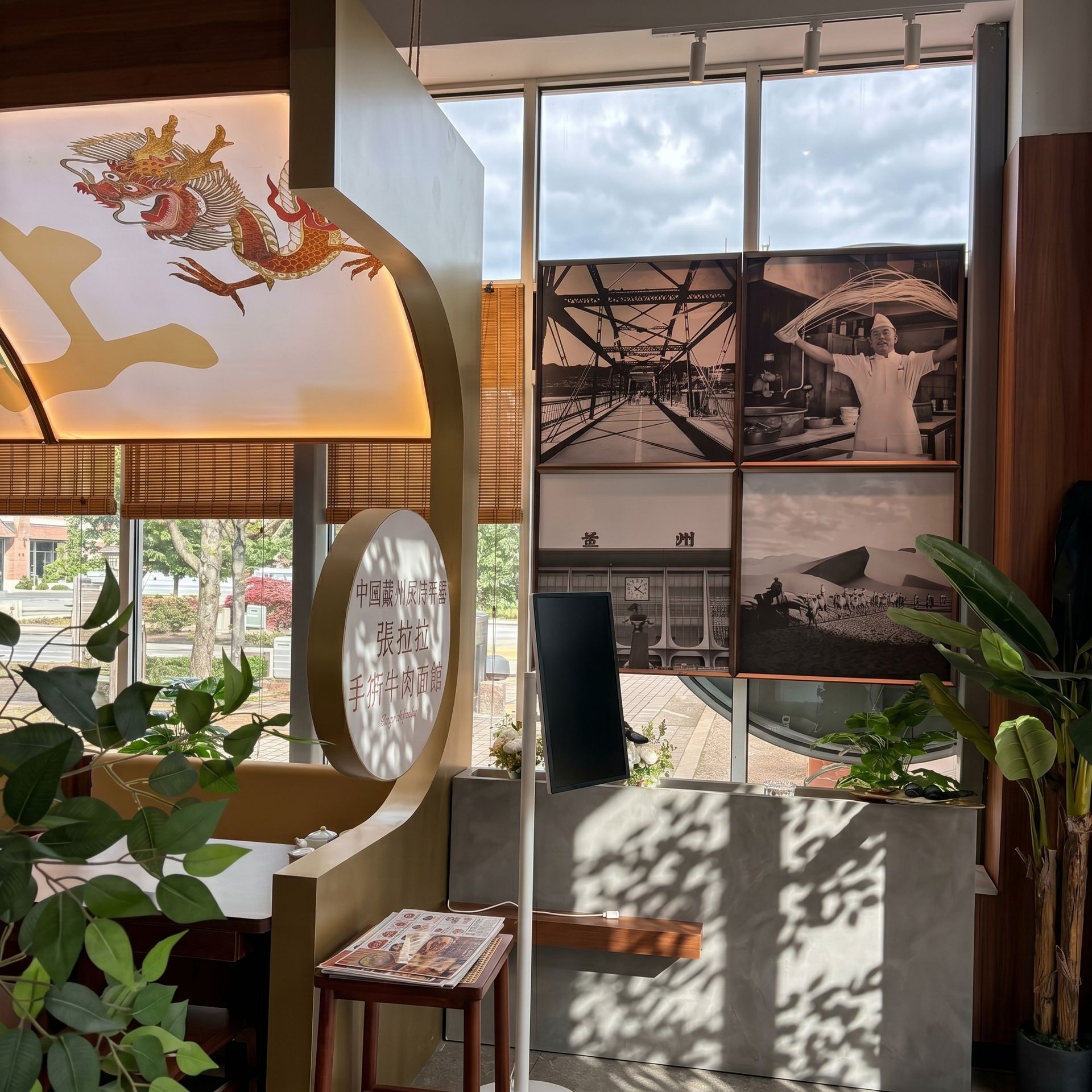 Sunlit café corner with plants, a wooden stool, and a wall of black-and-white travel photos