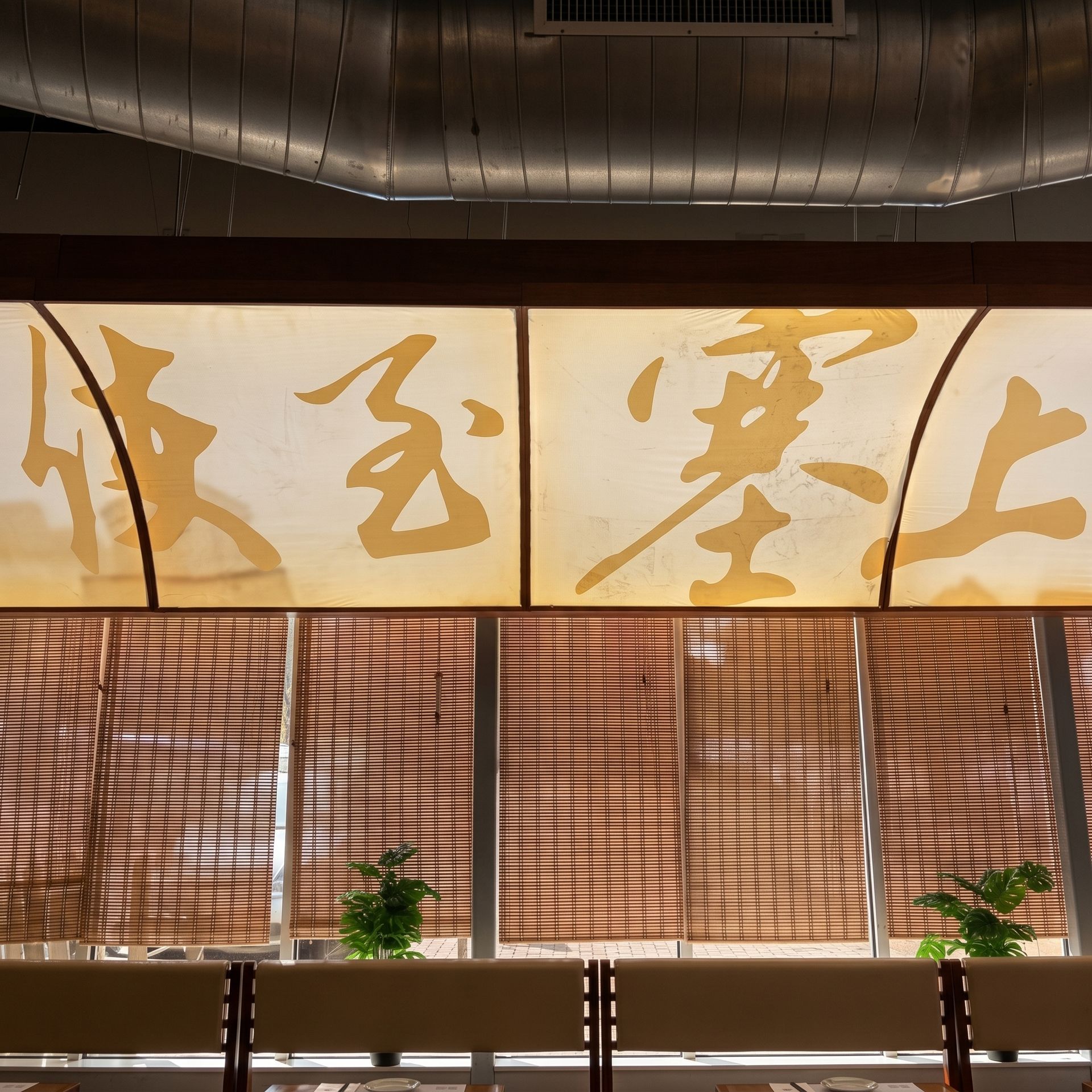 Interior with large gold calligraphy panels above bamboo blinds and potted plants.