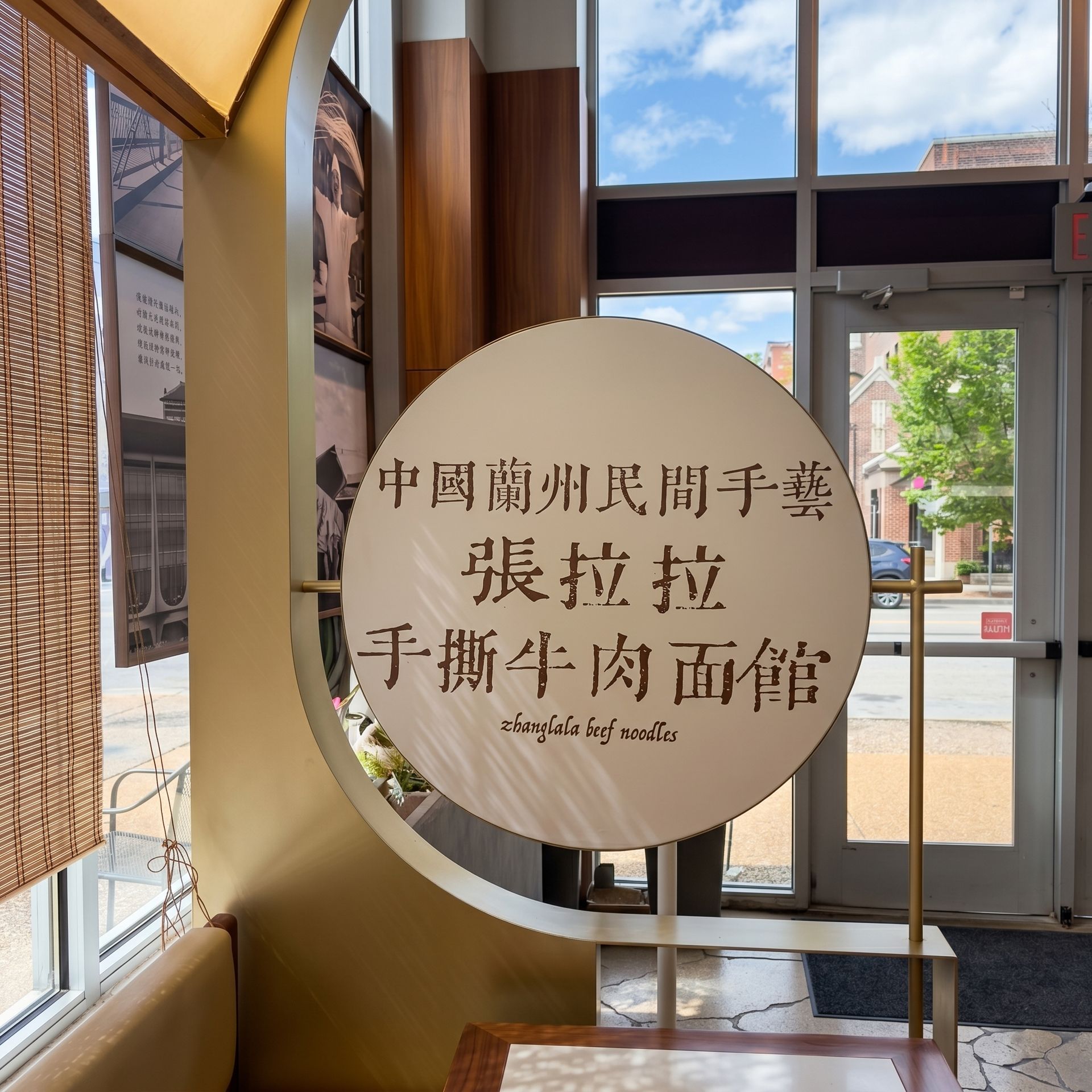 Round sign with Chinese text beside a sunlit glass entrance, with a poster board and outdoor view beyond.
