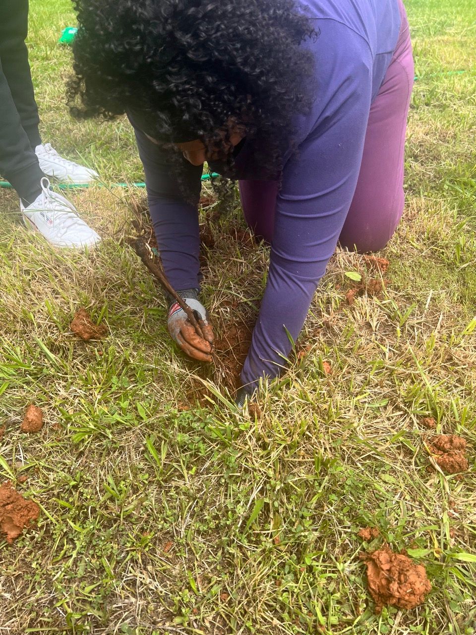 A woman is kneeling down in the grass holding a shovel.