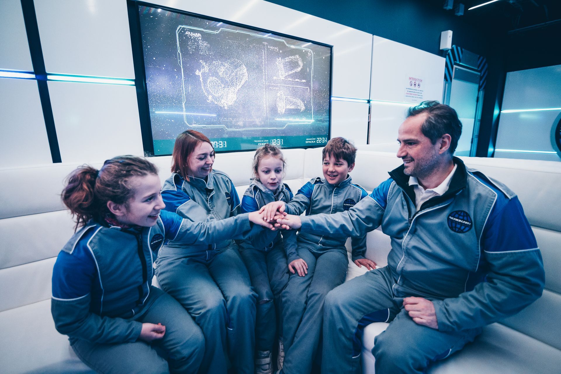 Un homme et quatre enfants en combinaison spatiale se blottissent ensemble dans une pièce futuriste.
