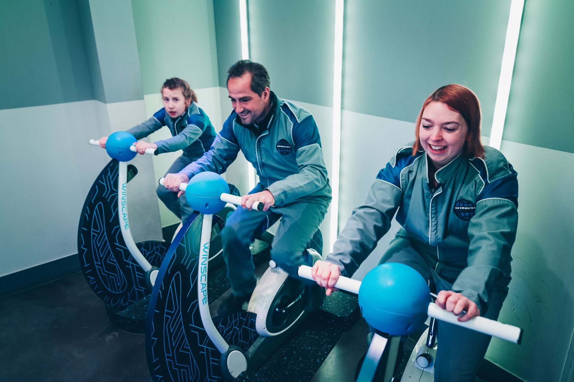 Trois personnes en combinaison spatiale font du vélo d'exercice dans une pièce d'escape game.