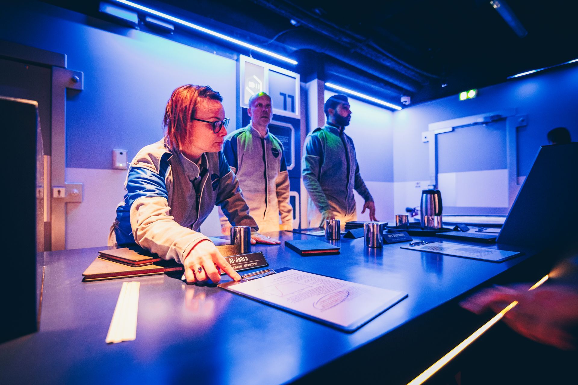 Trois personnes dans un décor futuriste. L'une d'elles pointe un presse-papiers ; éclairage bleu. Une salle d'escape game.