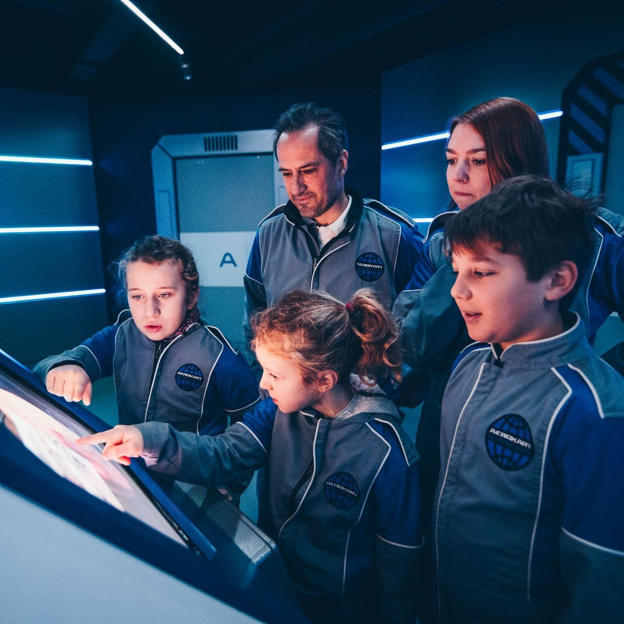 Une famille en combinaisons spatiales grises interagit avec un écran tactile dans une pièce éclairée en bleu.
