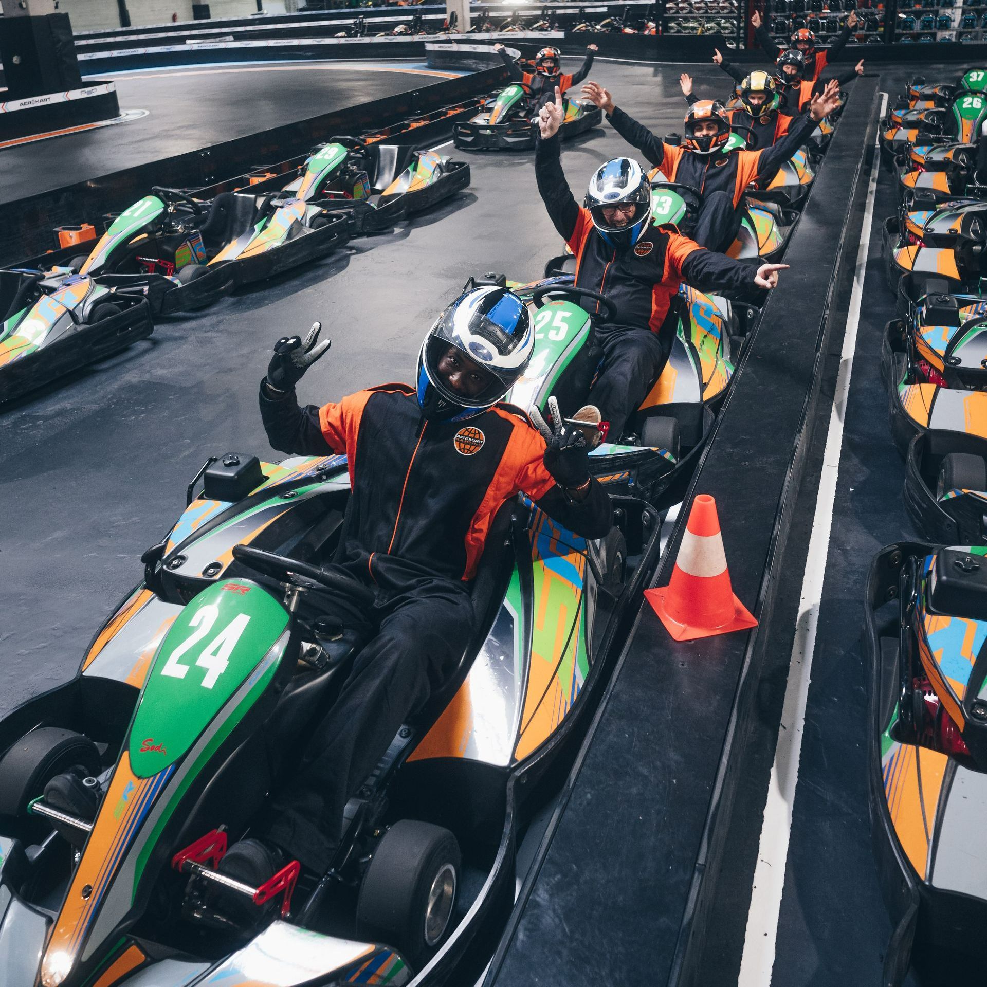 Des gens dans des karts levant les mains, portant des casques et des combinaisons, sur une piste de course intérieure.