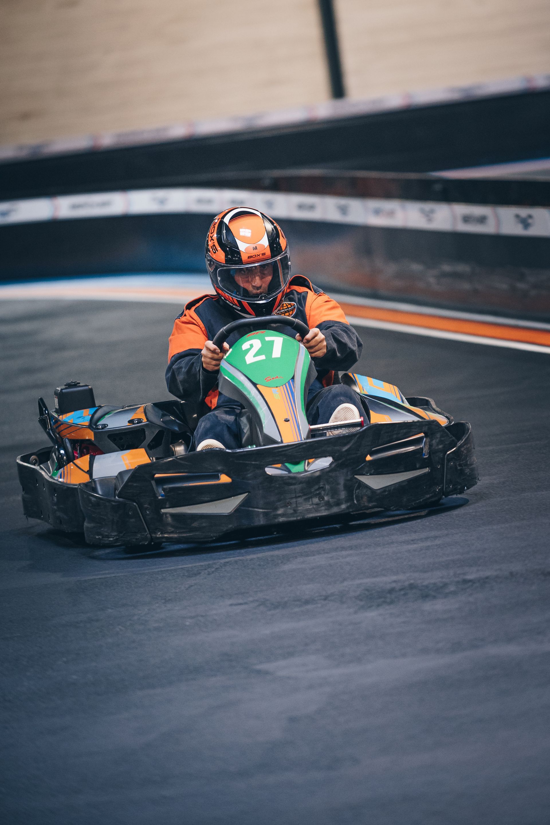 Un pilote de karting casqué, en pleine course sur une piste. Le kart est noir avec des touches d'orange, numéro 27.