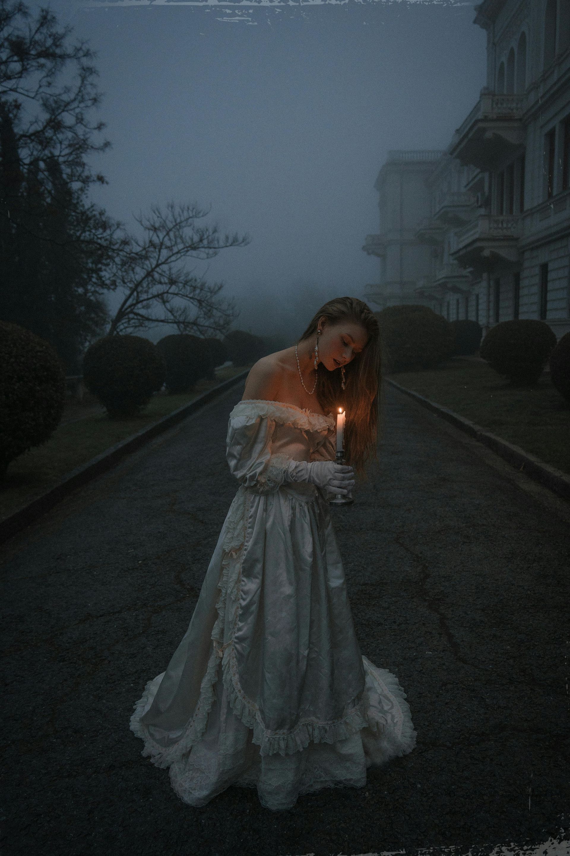Une femme en robe blanche tient une bougie sur un chemin brumeux devant une grande maison.