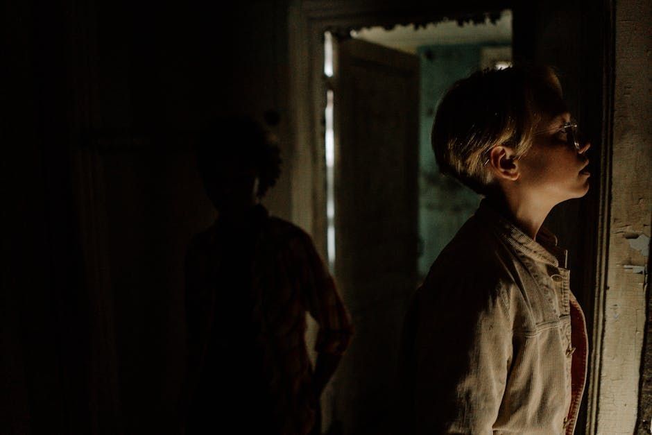 Child in light, looking through doorway. Another figure in the shadows. Dark, old interior.