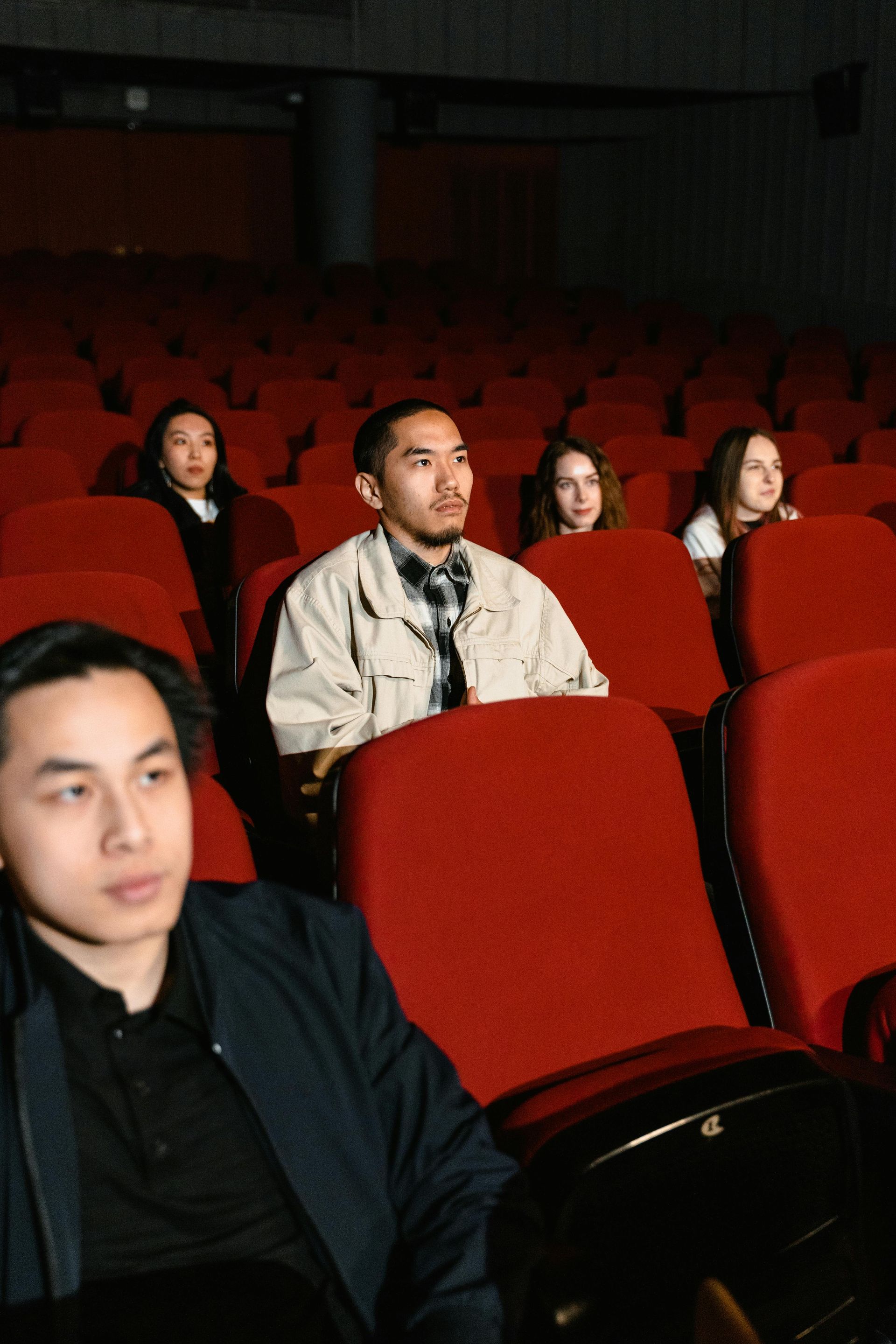 Cinq personnes assises dans des fauteuils de théâtre rouges, regardant quelque chose. Lumière tamisée.
