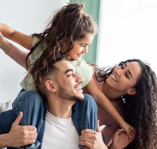 A man smiles, carrying a child on his shoulders while a woman smiles, standing nearby.