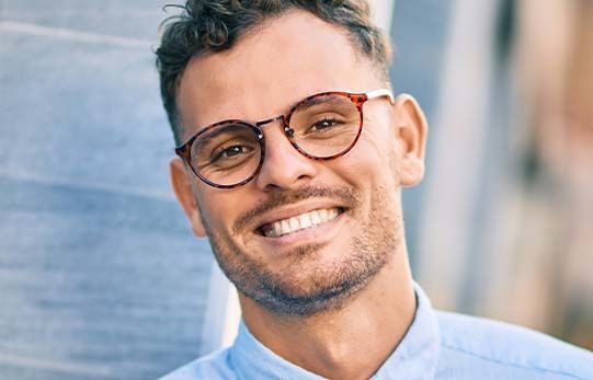 Man with glasses smiles brightly, light blue shirt, outside.
