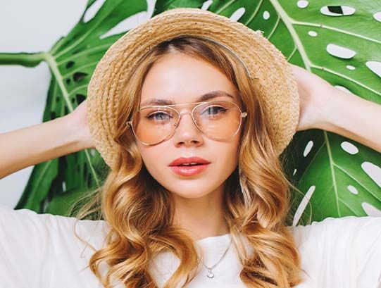 Woman in straw hat and glasses poses in front of large green leaf.
