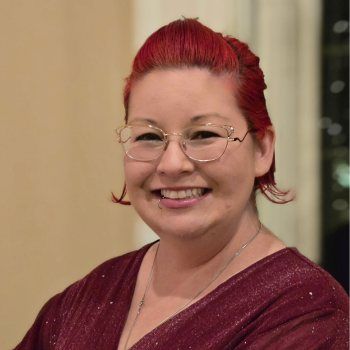 Dr. Christine Cosgrove with red hair and glasses smiles. Wearing a maroon top, indoor setting.