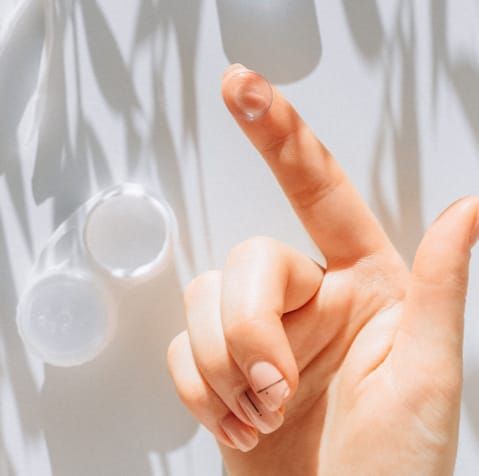 Hand holding a contact lens, with a lens case in the background, against a white surface with shadows.