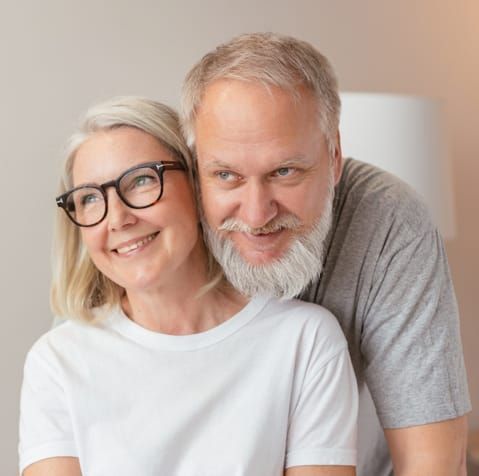 Woman wearing glasses smiles as man hugs her, both look happy, inside.