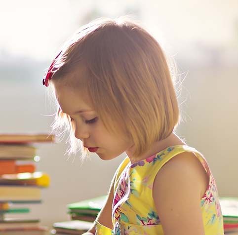 Girl with short blonde hair in a yellow floral dress, looking down at a book, with a stack of books in the background.