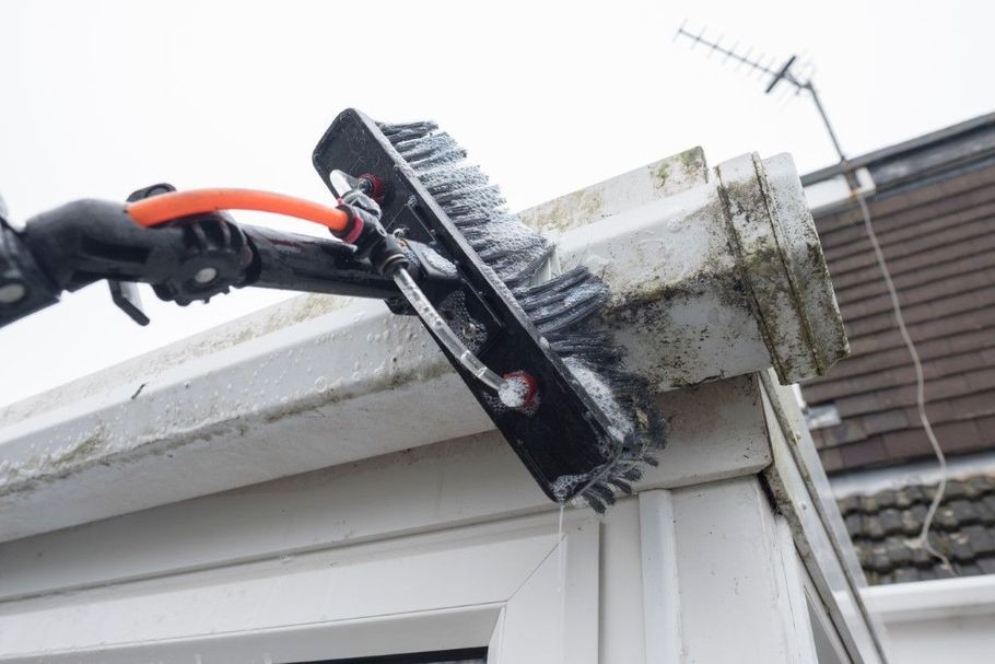 Brush cleaning a white window frame and gutter edge outdoors with a long-handled tool.