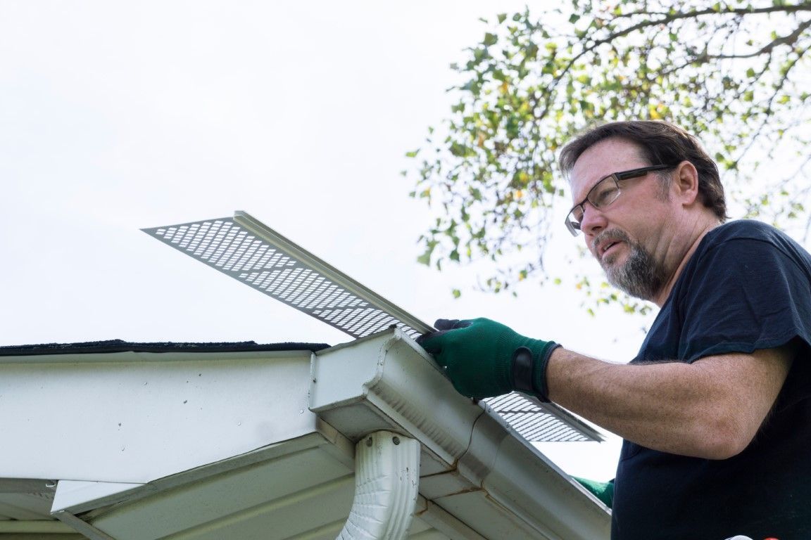 Man wearing gloves cleaning a white gutter on a roof with a metal brush tool