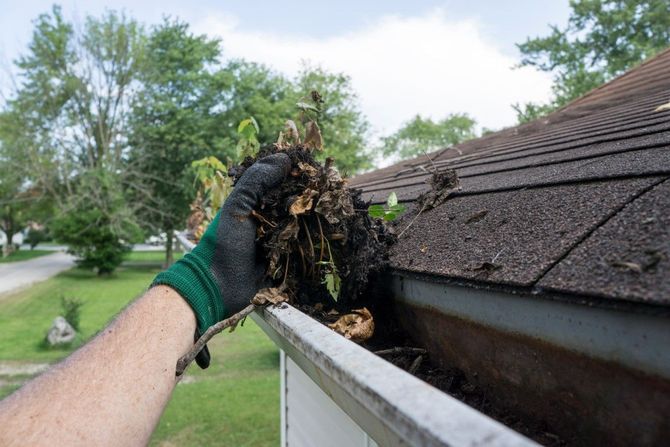 Gloved hand removing debris from a gutter beside a shingled roof on a suburban home