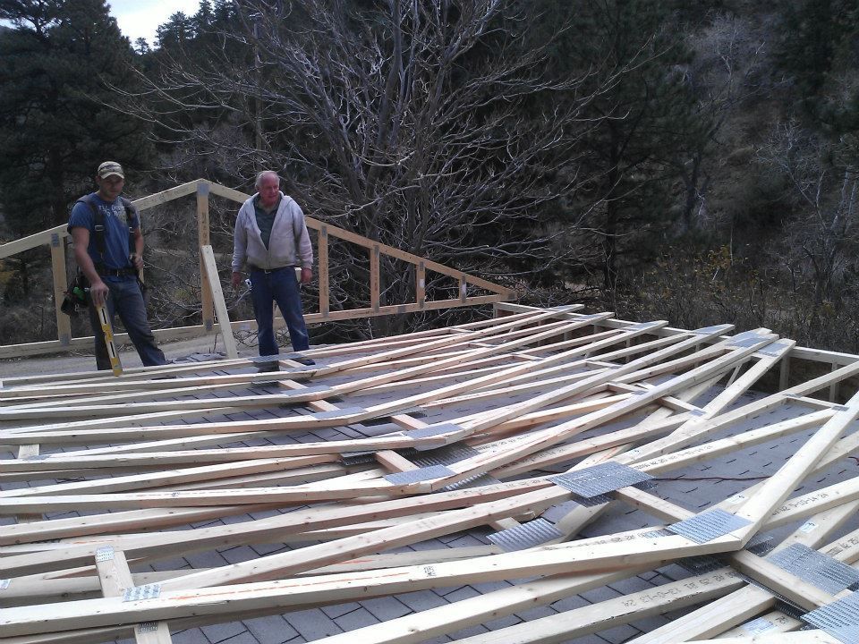 Two workers inspecting a partially built wooden roof frame outdoors in a wooded area