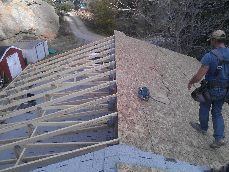Roof framing and sheathing under construction, with a worker on a sloped roof and a dirt path in the background.