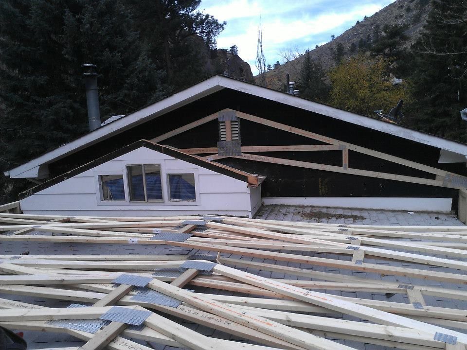 House under construction with roof trusses and lumber piles in front, set against a mountain backdrop