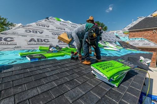 Worker on a roof installing shingles, with stacks of green roofing materials against a blue sky.