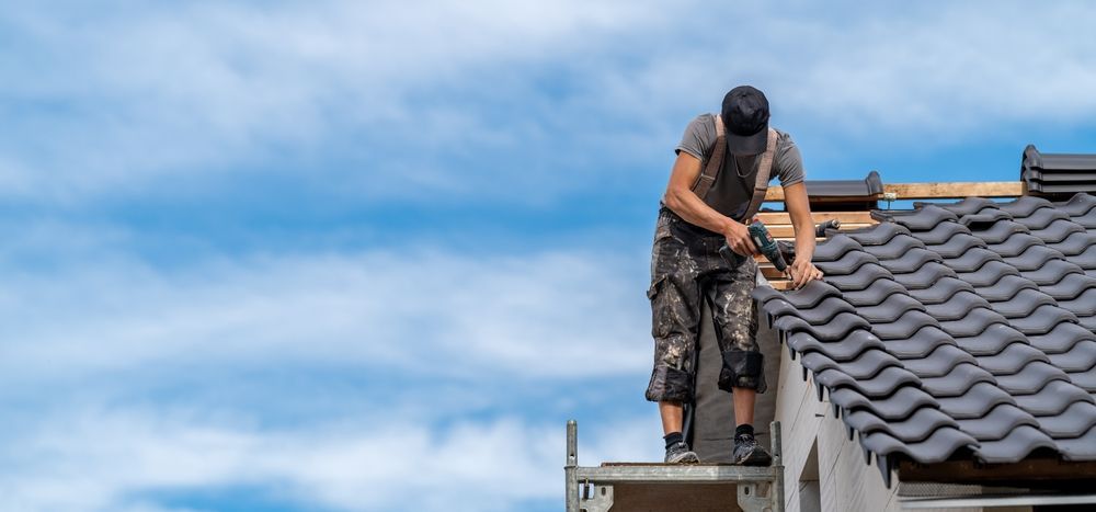 Roofing worker kneels on a tiled roof under a blue sky, adjusting tiles near the edge.