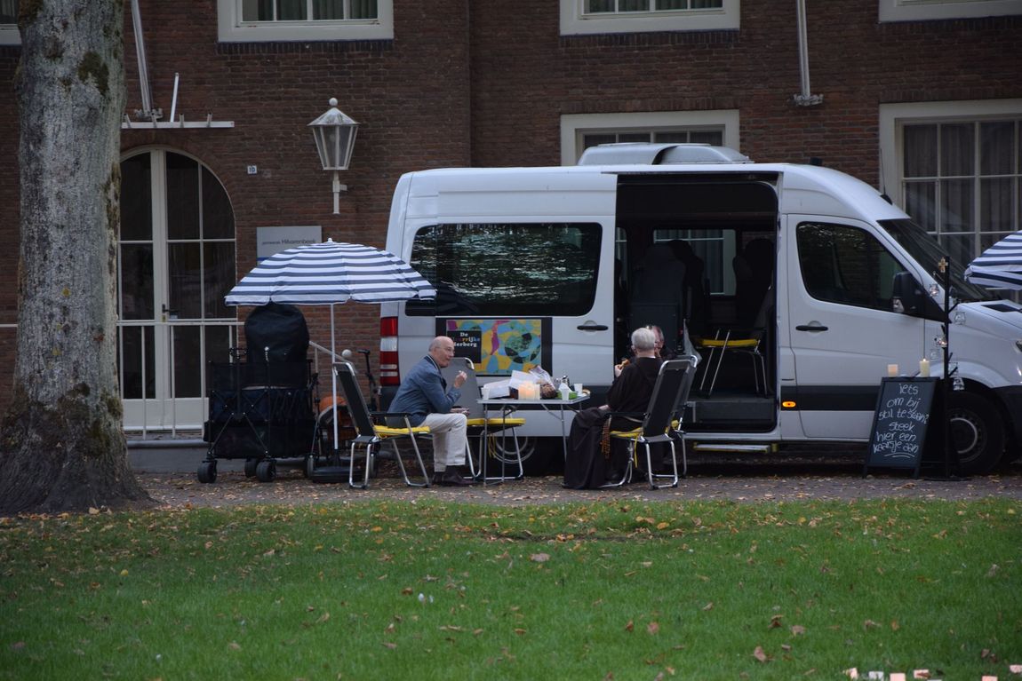 Mensen zitten aan een tafel naast een witte bestelwagen op een grasveld, met een parasol en een bakstenen gebouw op de achtergrond.