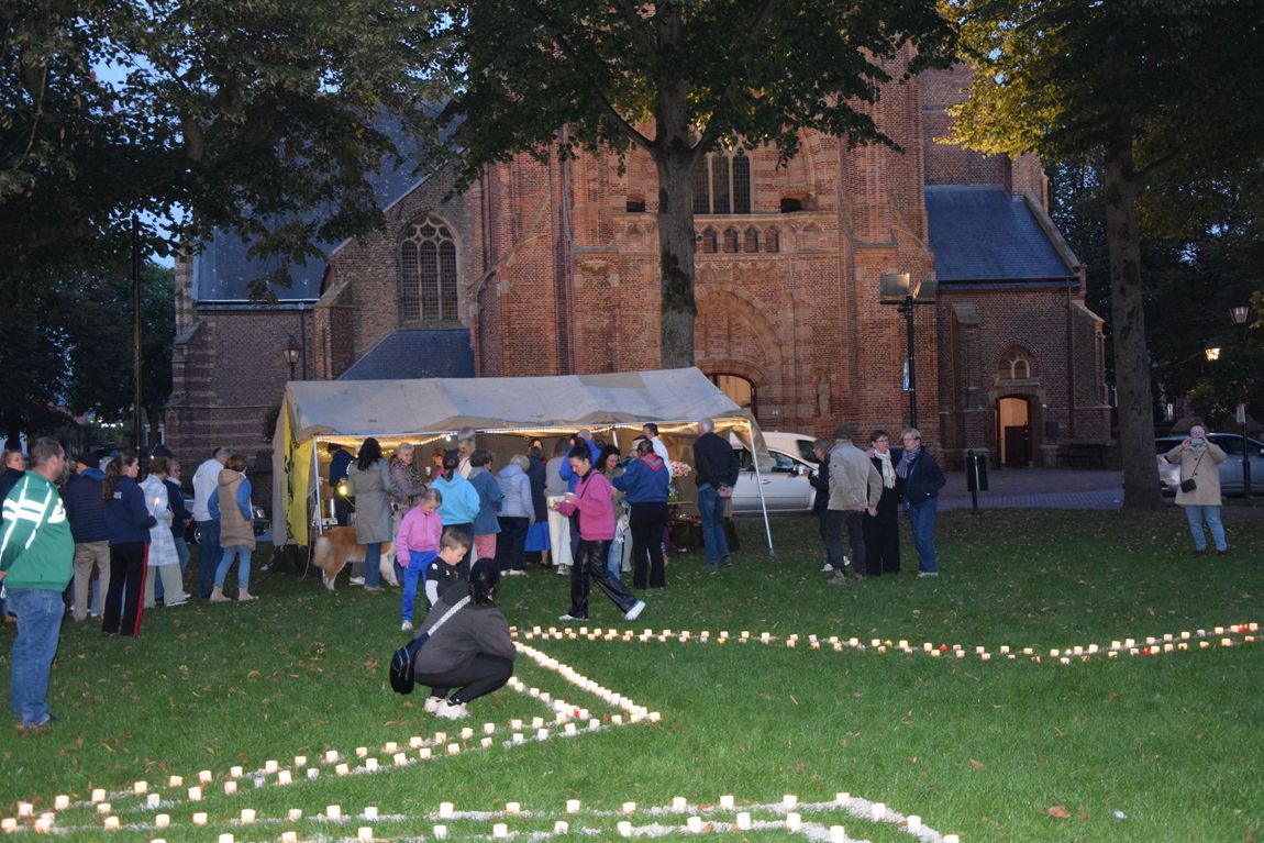 Mensen verzamelden zich bij schemering op een grasveld naast een verlichte tent en een bakstenen gebouw, waar kaarsen in cirkels waren opgesteld.