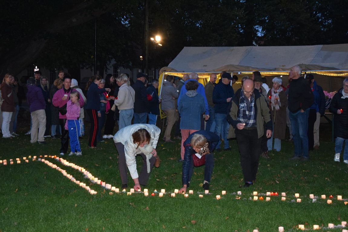 Mensen steken kaarsen aan op een grasveld in de schemering, met een menigte verzameld onder een tent achter hen.