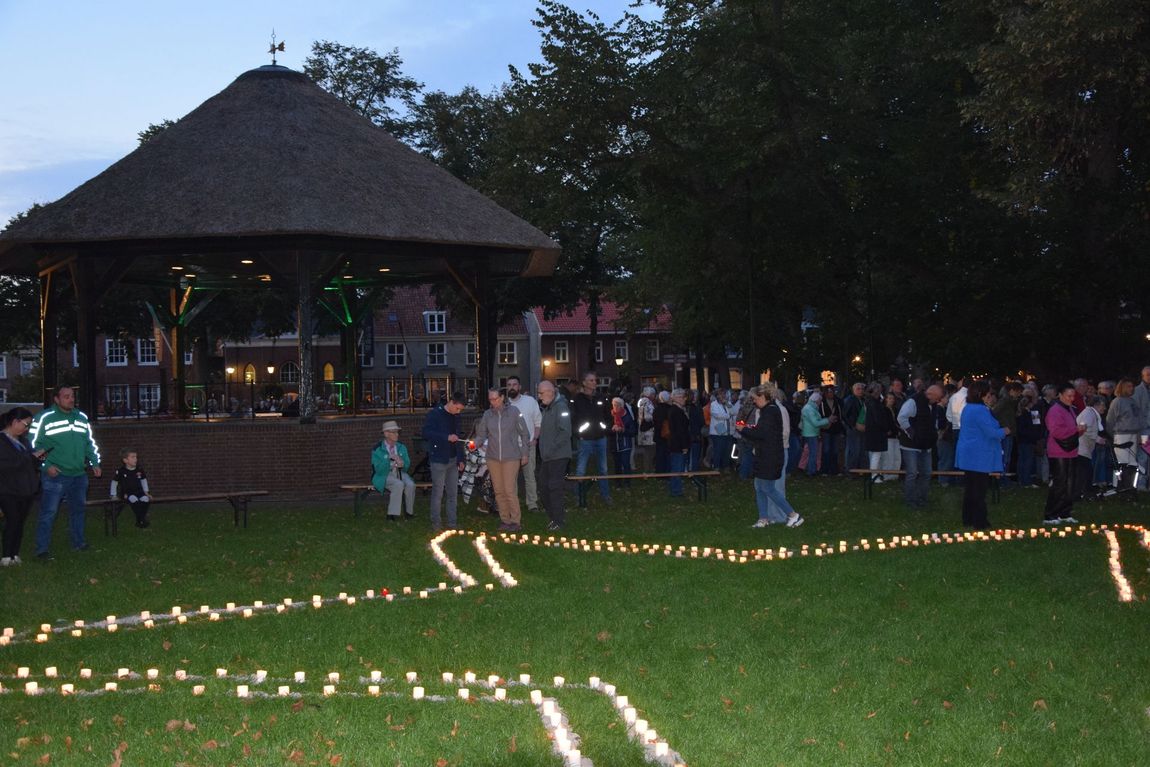 Mensen verzamelden zich bij schemering rond een verlicht paviljoen, waar kaarsen de paden over een grasveld markeerden.